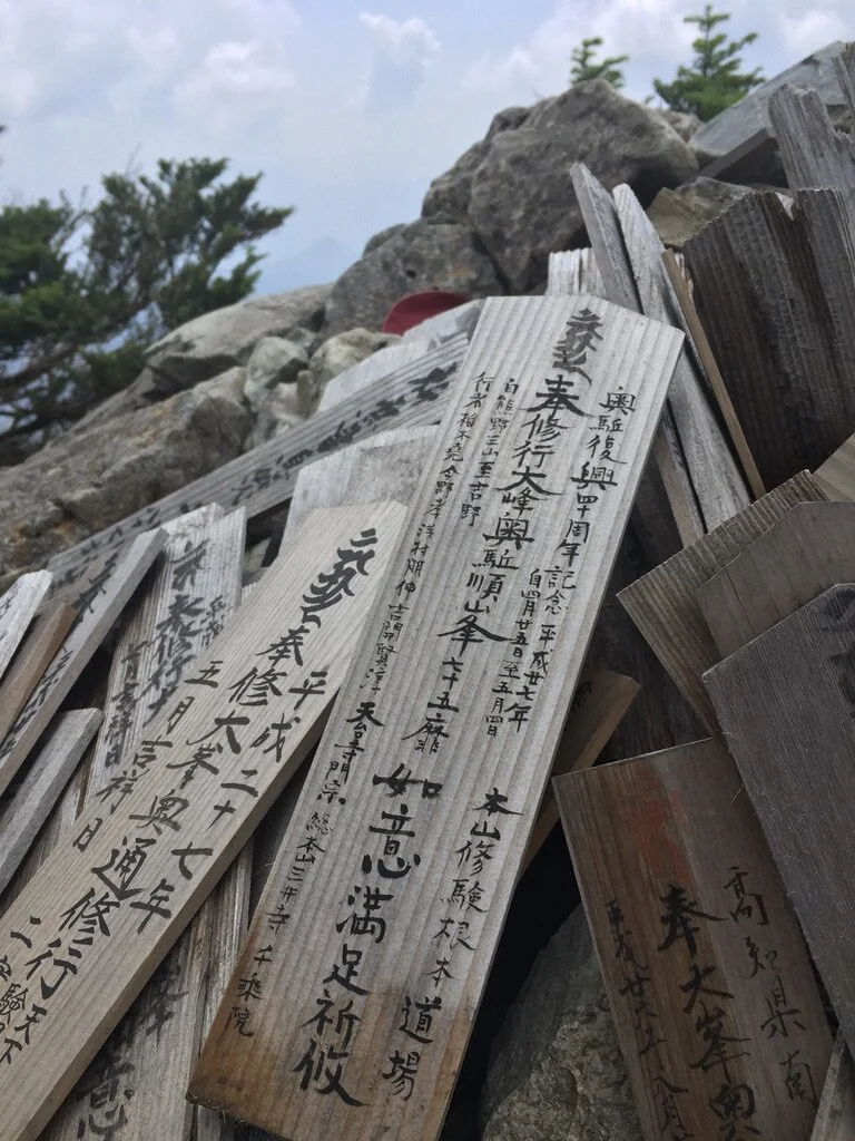Multiple wooden prayer plaques with Japanese writing attached to a rock and surrounded by trees under a cloudy sky.