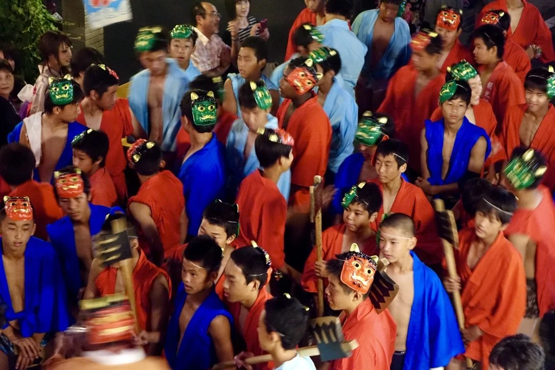 Group of young boys dressed in traditional Japanese festival attire, some wearing colorful masks, participating in a cultural festival or event.