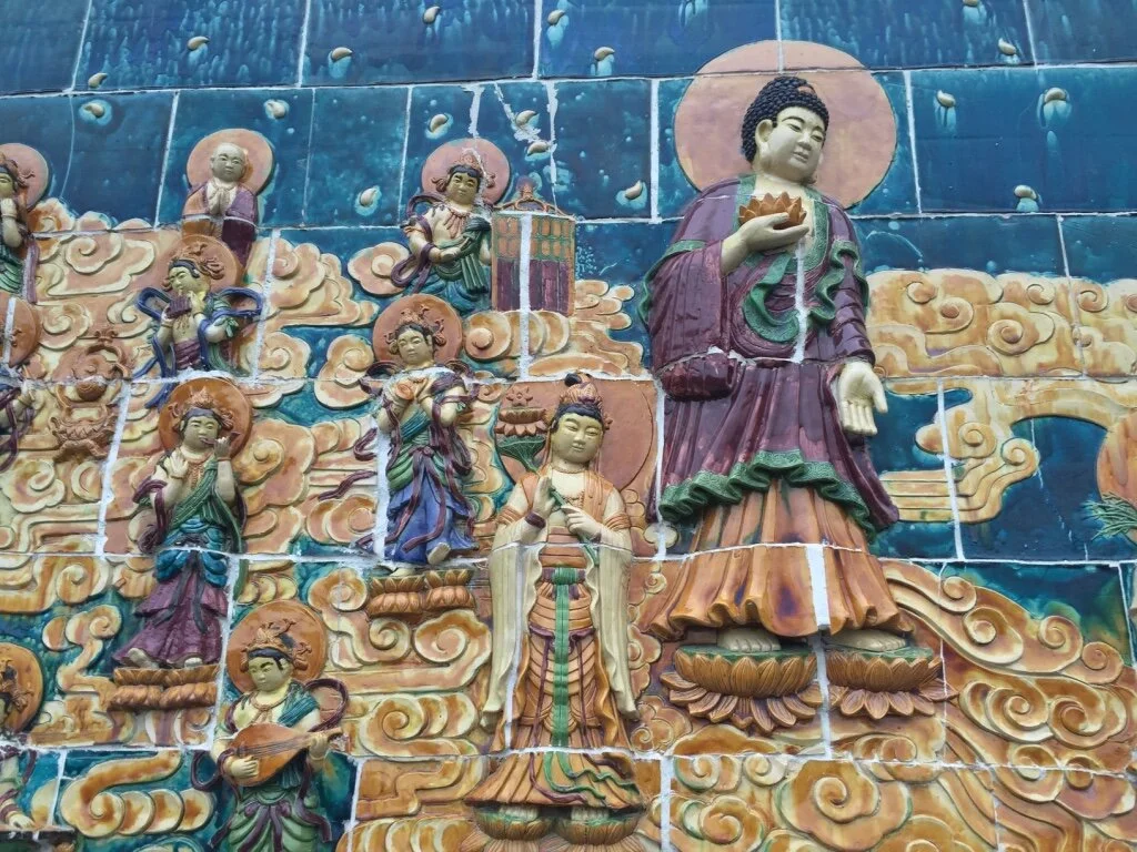 Colorful ceramic relief of Buddhist figures on a blue cloud background, depicting a large standing figure with a halo and smaller figures surrounding it.