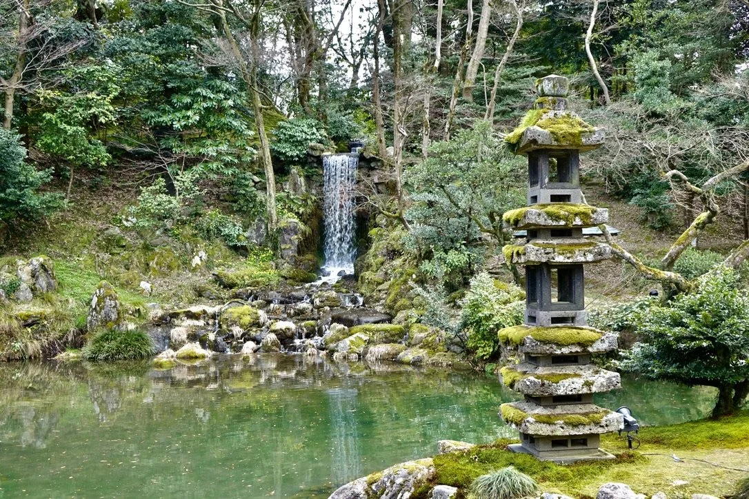 A lush garden scene with a pond, moss-covered stone pagoda on the right, and a waterfall in the background surrounded by trees and greenery.