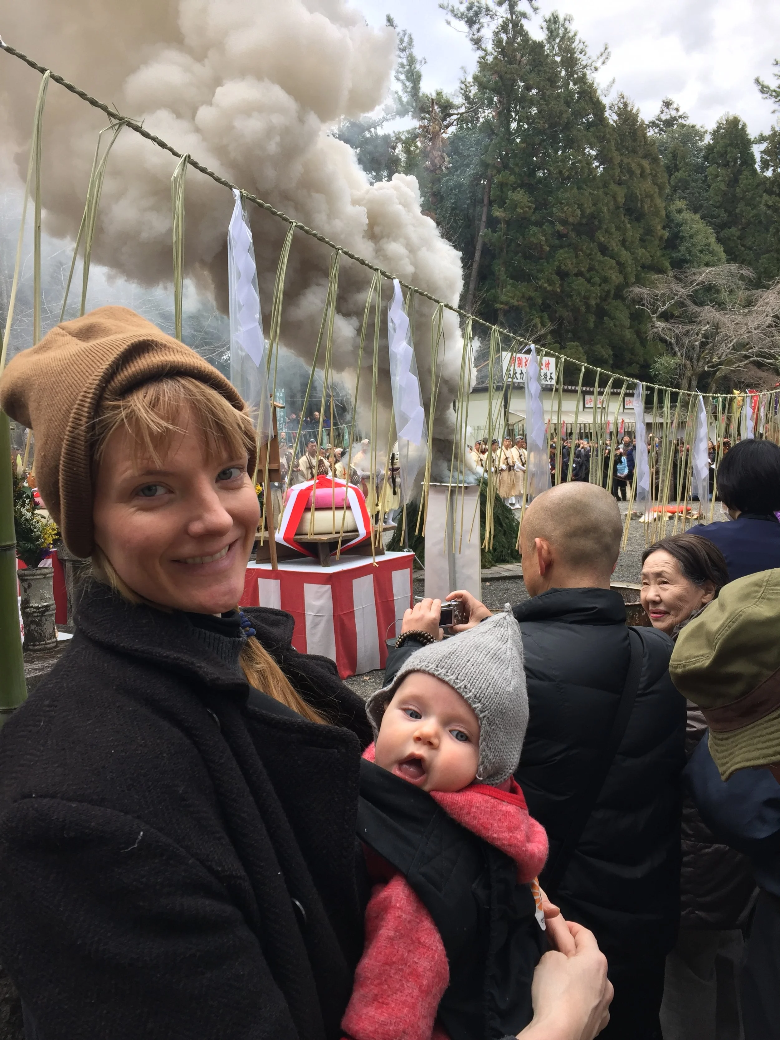A woman holding a baby at a traditional Japanese festival, with a large cloud of white smoke in the background, people gathering, and decorative paper streamers hanging from a rope.