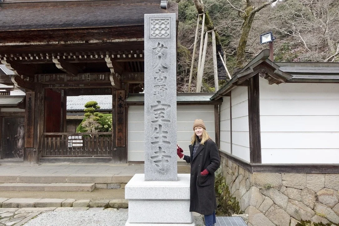 A woman standing next to a large stone monument with Japanese inscriptions in front of a traditional Japanese temple entrance.