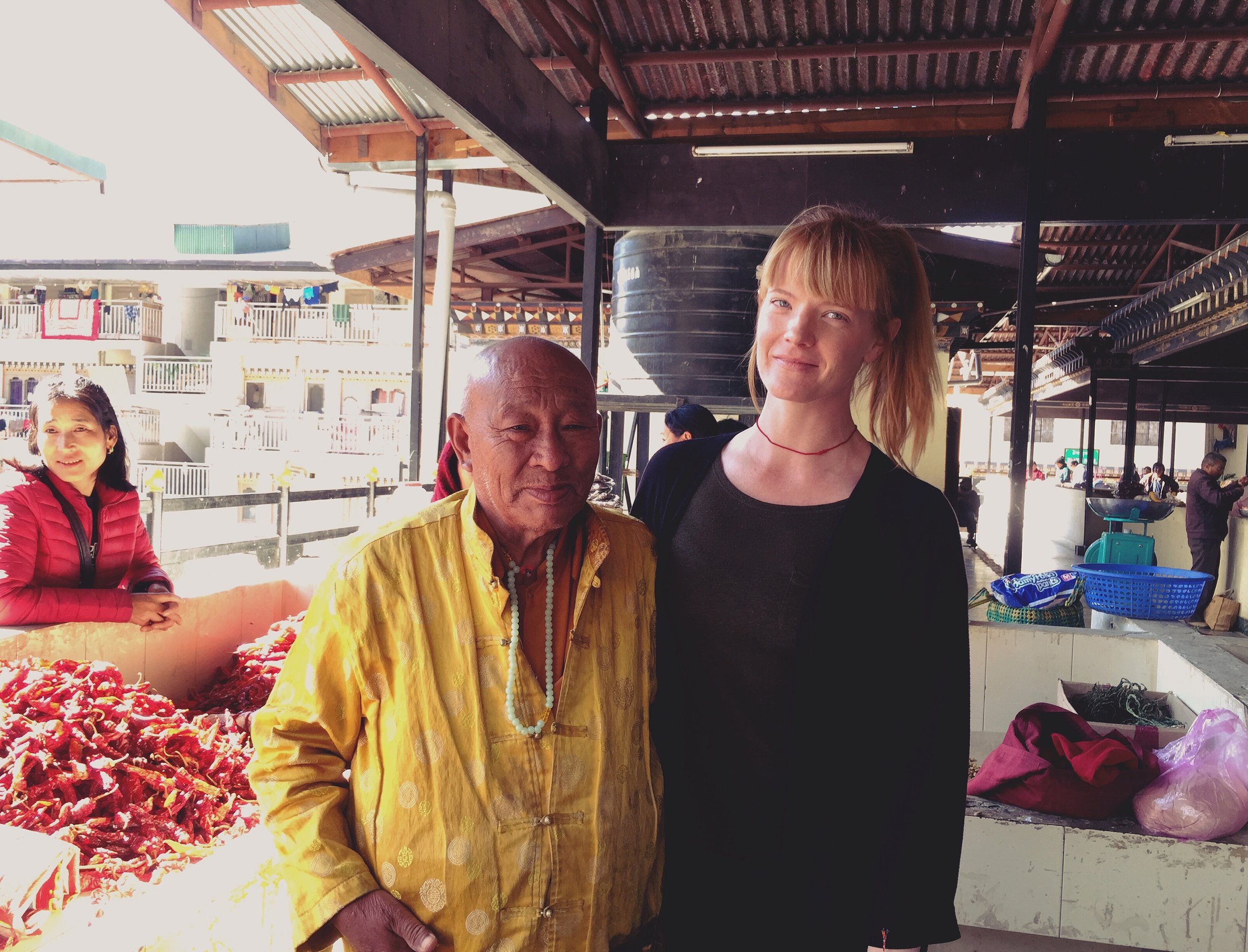 A young woman with blonde hair wearing a black top and a red necklace standing next to an elderly man in yellow traditional attire and beaded necklaces at an outdoor market. A woman in a red jacket is visible in the background, with market stalls and
