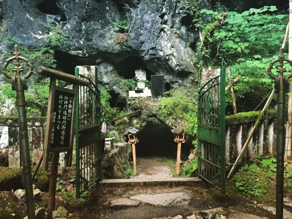 Open metal gate leading to a small cave or grotto in a rocky hillside, surrounded by green foliage and moss-covered rocks, with Japanese signs nearby.