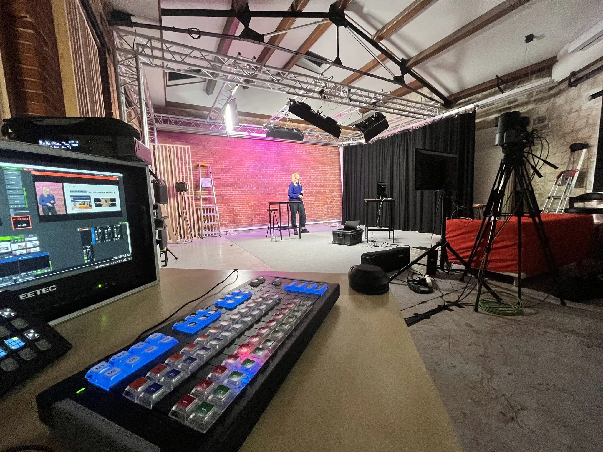 A woman is on a stage in a studio or event space with a brick wall background. The scene includes lighting and camera equipment, as well as a control panel in the foreground.