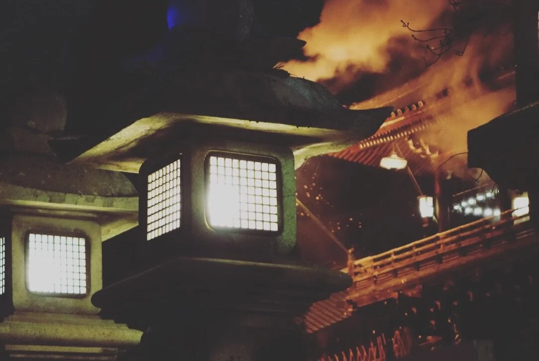A fire burning at night near traditional Japanese lanterns and a temple roof.