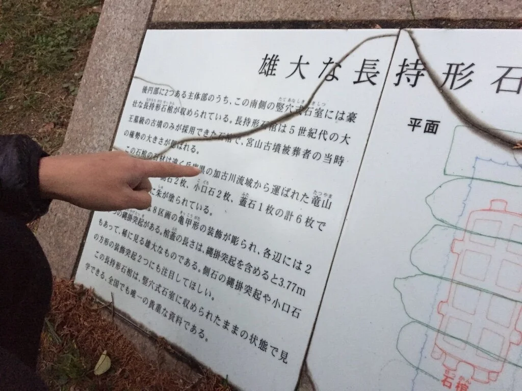 Person pointing at an informational sign in Japanese with a hand and part of the person's jacket visible, located outdoors near grass and concrete pavement.