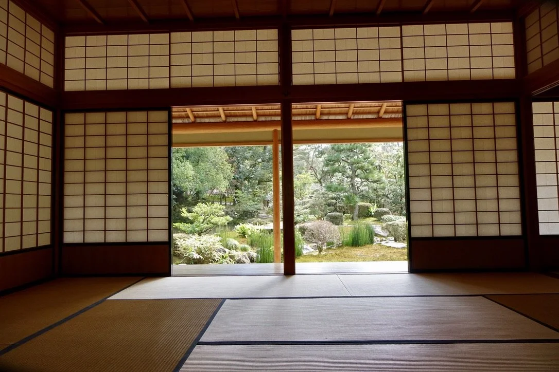 Traditional Japanese tatami room with sliding shoji doors open to a garden with rocks, trees, and plants.