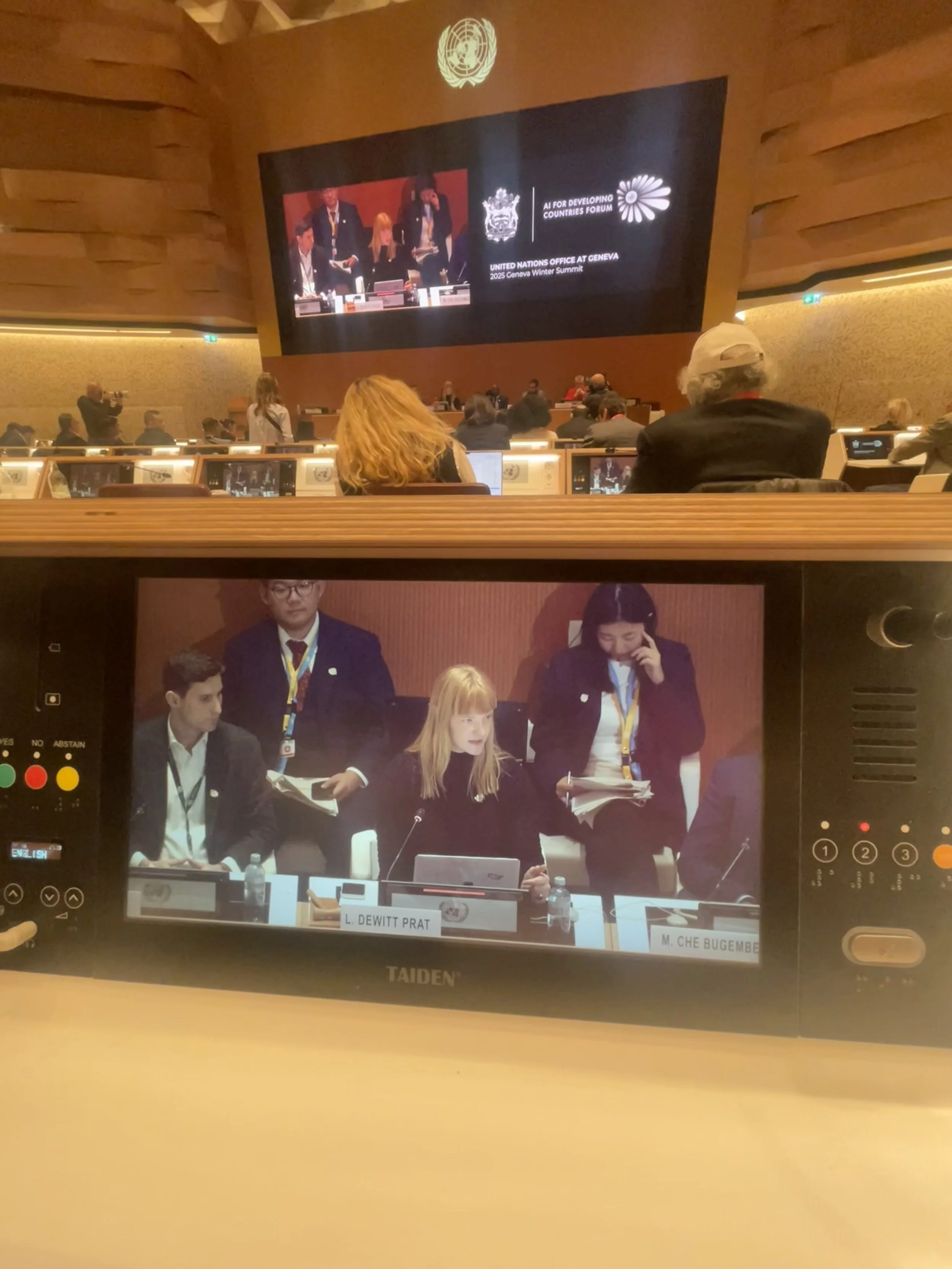 A view of a conference room showing people seated and a large screen displaying a United Nations event at Geneva Winter Summit, with the focus on a camera monitor capturing a panel including a woman in black sitting at a desk with a nameplate reading