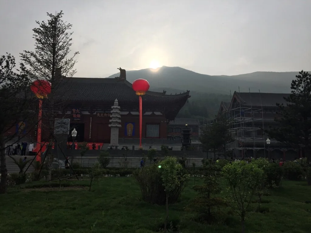 Traditional Asian temple with lanterns, pagoda, and scaffolding in front, set against a mountainous background with the sun partially hidden behind the mountains.
