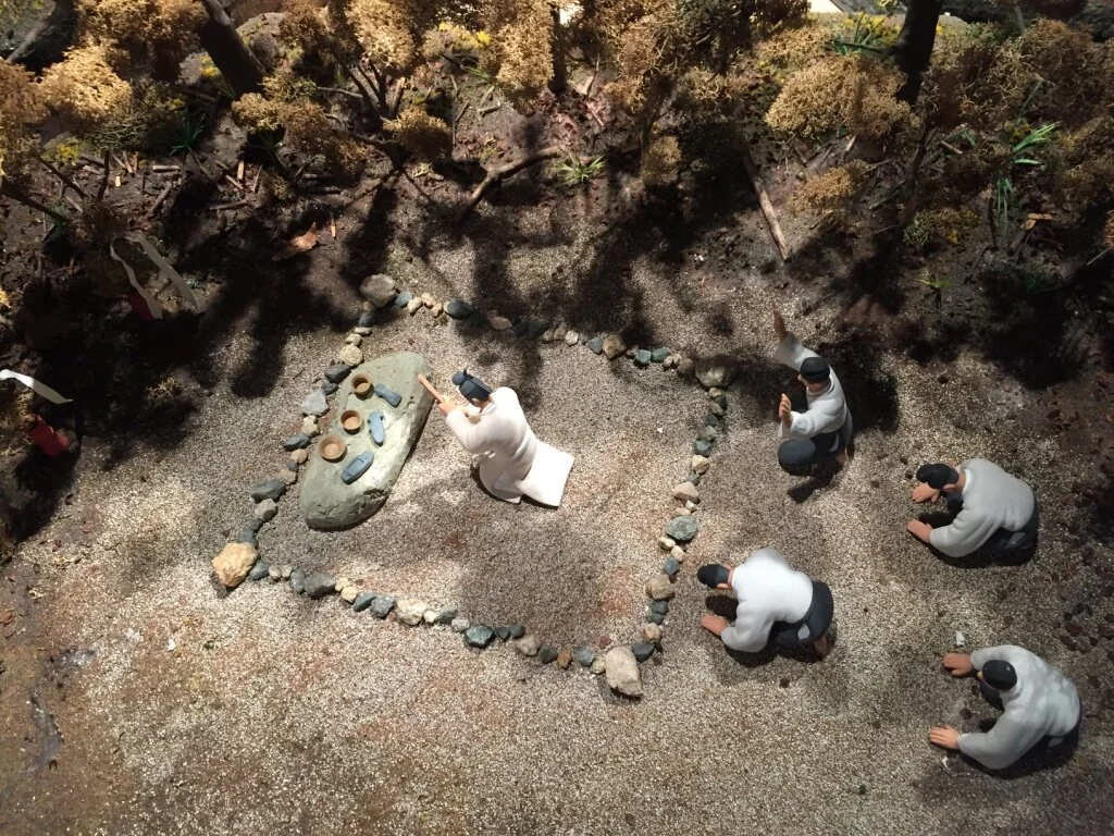 Statues of five people praying or paying respect around a heart-shaped arrangement of rocks with a large stone in the center, set outdoors with trees and bushes in the background.