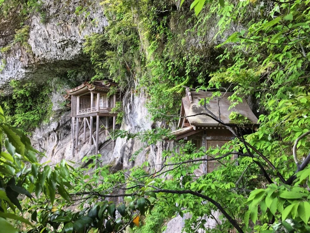Wooden Japanese-style shrine and small building built into a rocky cliffside surrounded by green foliage.