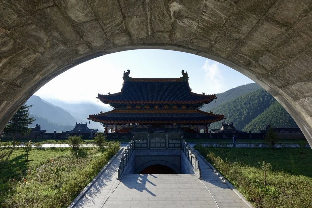 Traditional Chinese-style building seen through an archway, with mountains in the background and a landscaped pathway leading to the entrance.