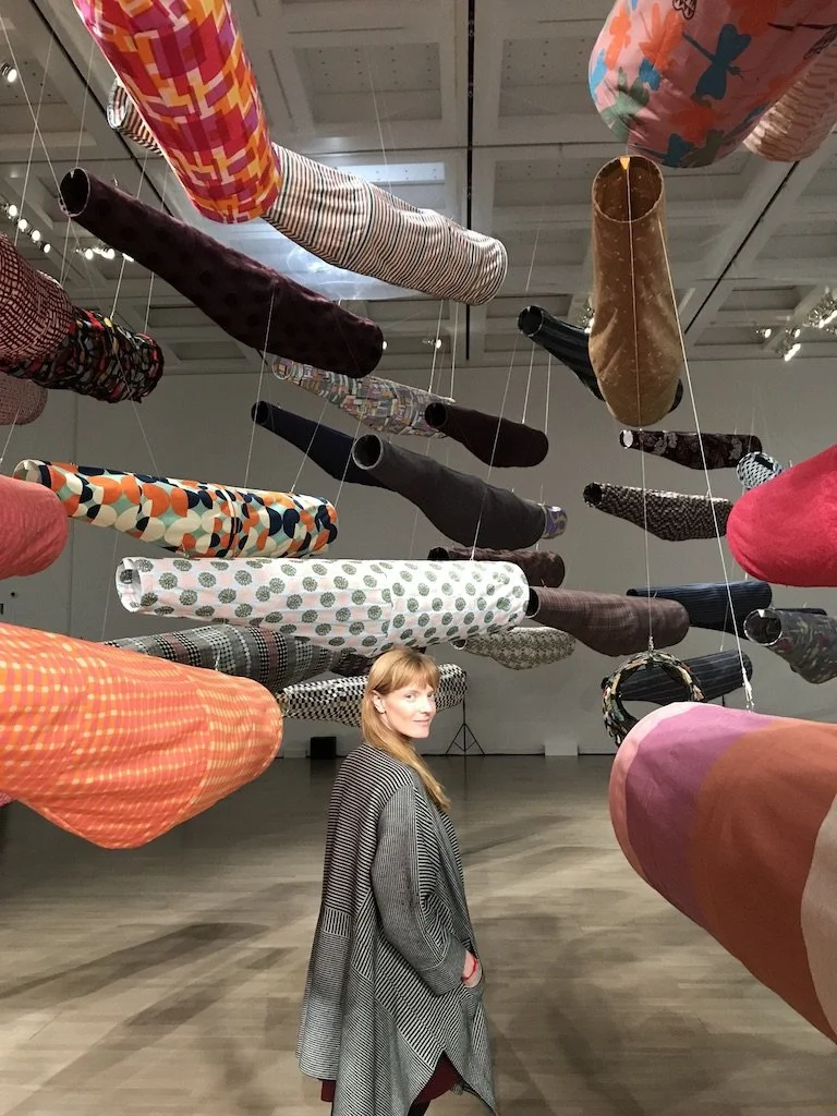 A woman with red hair wearing a checkered jacket stands in an art installation with colorful fabric tubes hanging from the ceiling in a gallery.