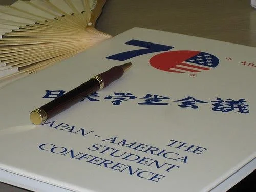 A brochure for the Japan-American Student Conference 70th anniversary, with a fountain pen placed on top, and a traditional folding fan in the background.