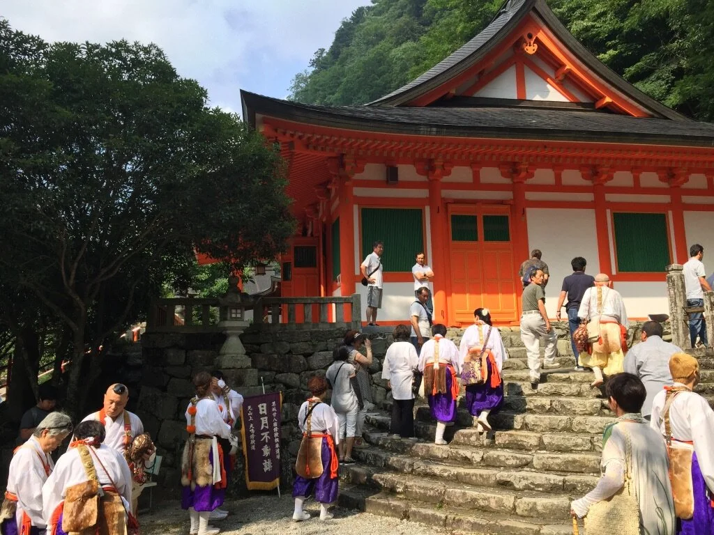 People dressed in traditional attire walking up stone steps toward a Japanese shrine building painted in orange and white, surrounded by greenery.