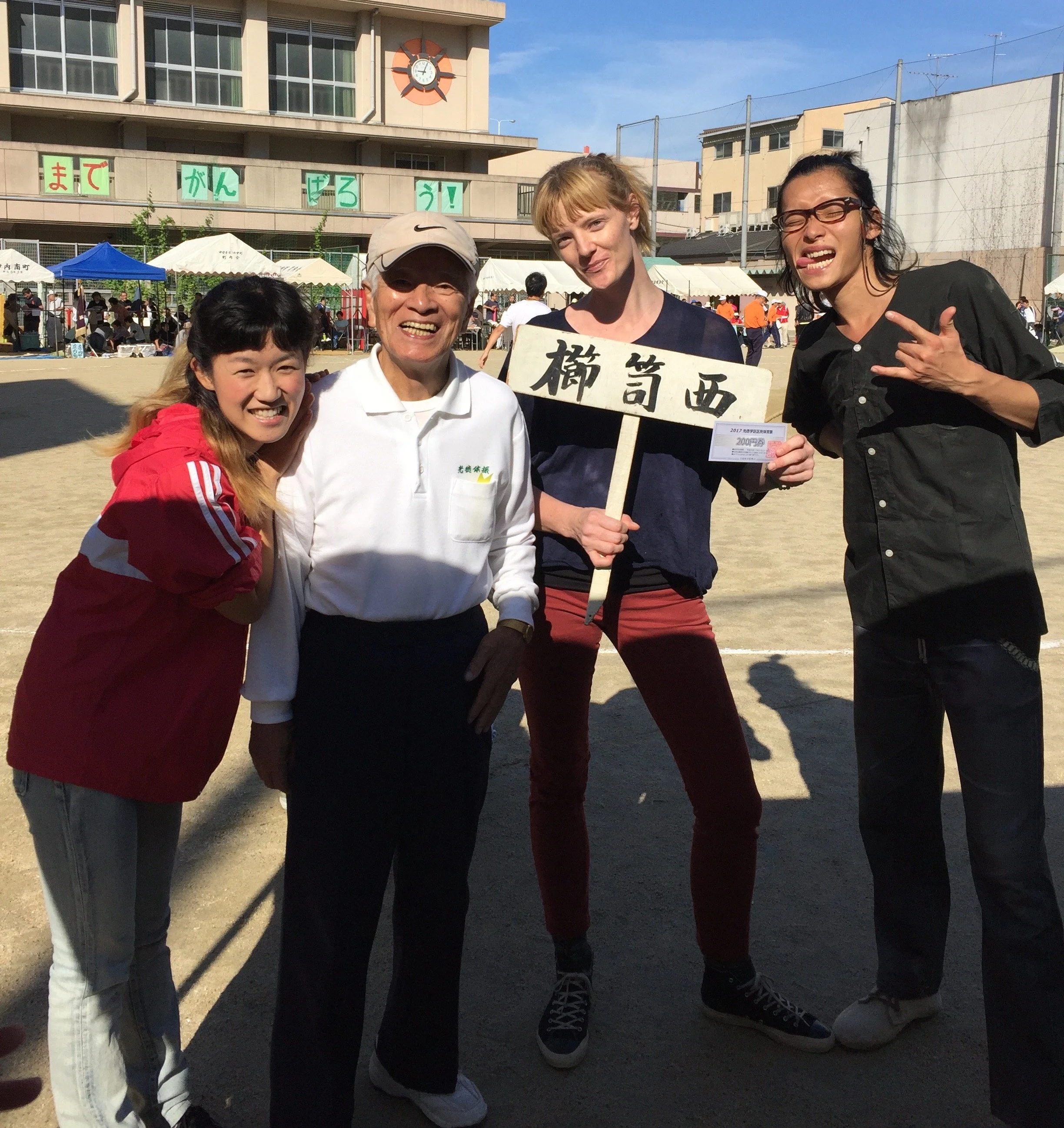 Four people are standing outdoors on a sunny day, smiling for the photo. They are in front of a school or community building with a clock and tents, possibly during a festival or outdoor event. One woman is holding a sign with Japanese characters.