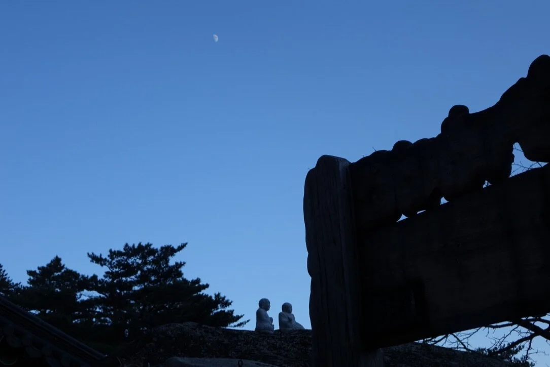 Silhouette of two statues and a carved wooden beam against a dusk sky with the moon visible in the distance.