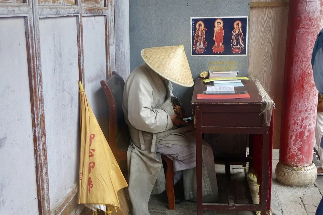 Person wearing traditional Asian conical hat sitting at a small wooden table using a mobile phone, with a Buddhist poster on the wall behind, an umbrella hanging on the wall, and a red pillar to the right.