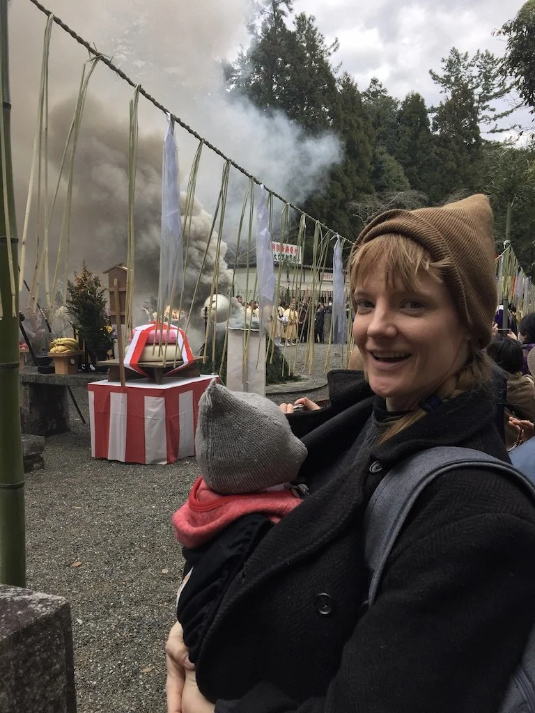 A woman smiling while holding a child with a gray hat, at a festival or outdoor event with smoke and people in the background.