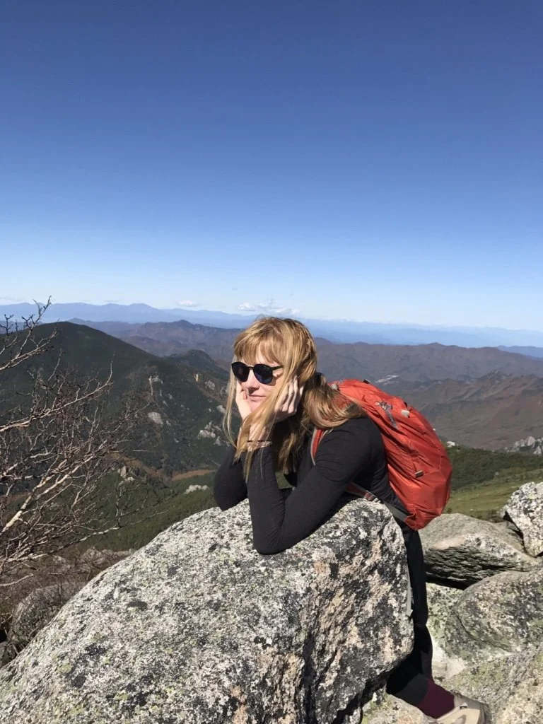 A woman with blonde hair wearing sunglasses, a black long sleeve shirt, and a red backpack, leaning on a large rock on a mountain trail, with a scenic view of mountains and a blue sky in the background.