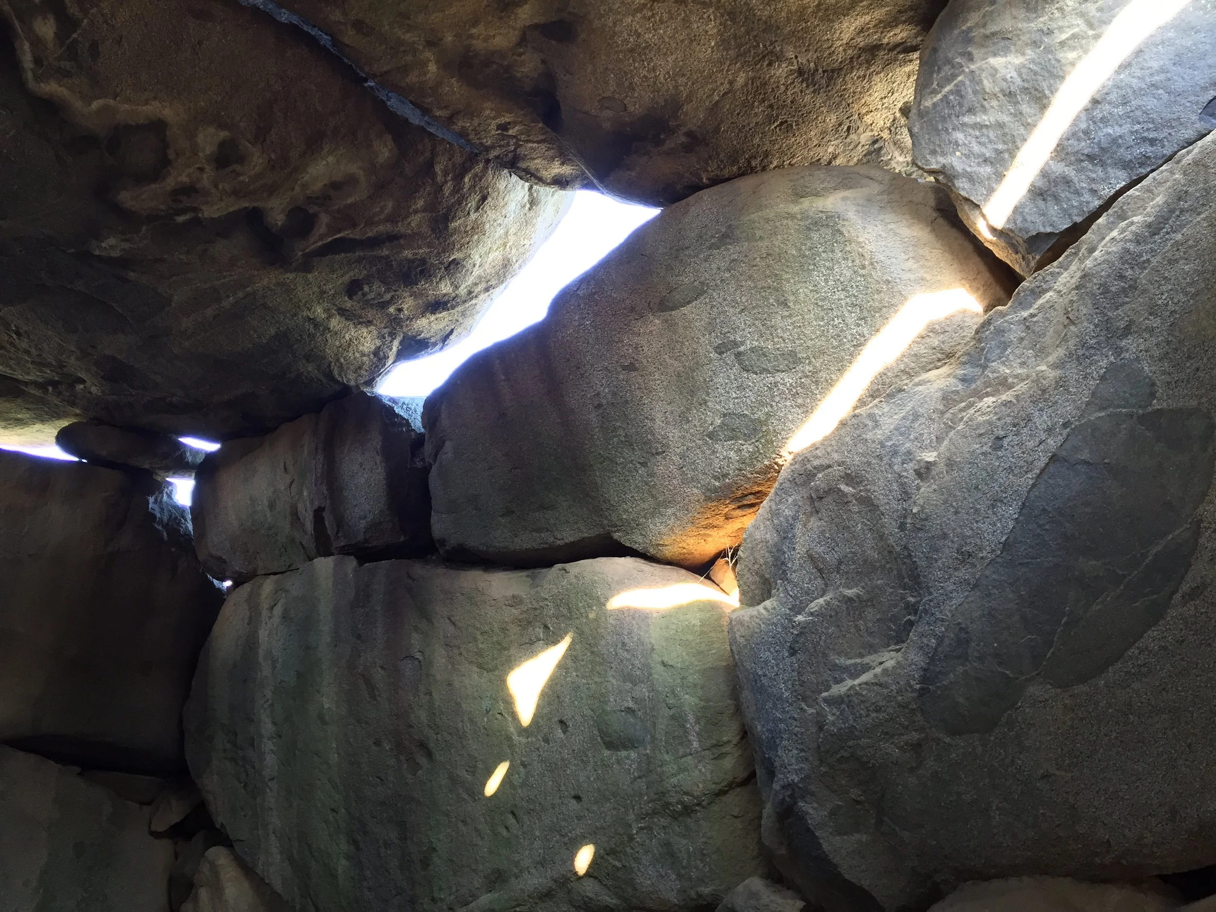 A small opening between large, rough rocks, with sunlight streaming through and creating bright patches on the surface of the rocks.