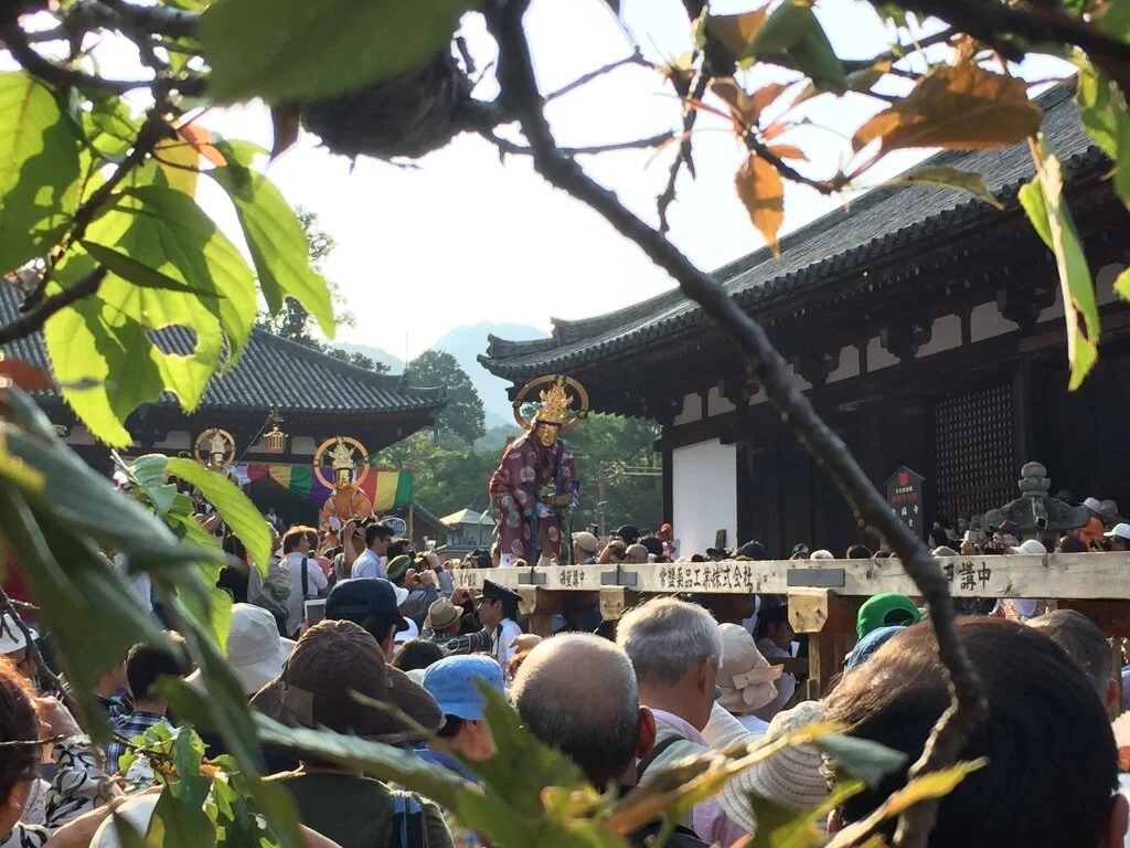 Crowd attending a traditional outdoor event with statues of deities on a wooden platform in front of a temple, surrounded by trees and foliage.