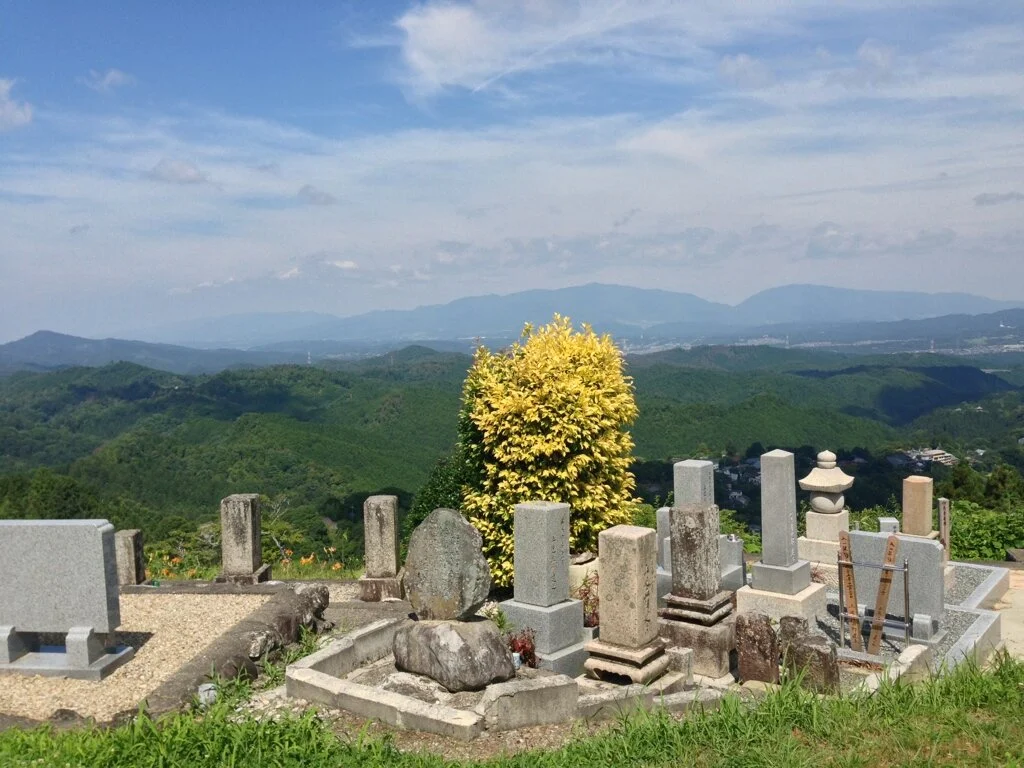 A rural cemetery with several gravestones and monuments, overlooking green, hilly mountains under a partly cloudy sky.