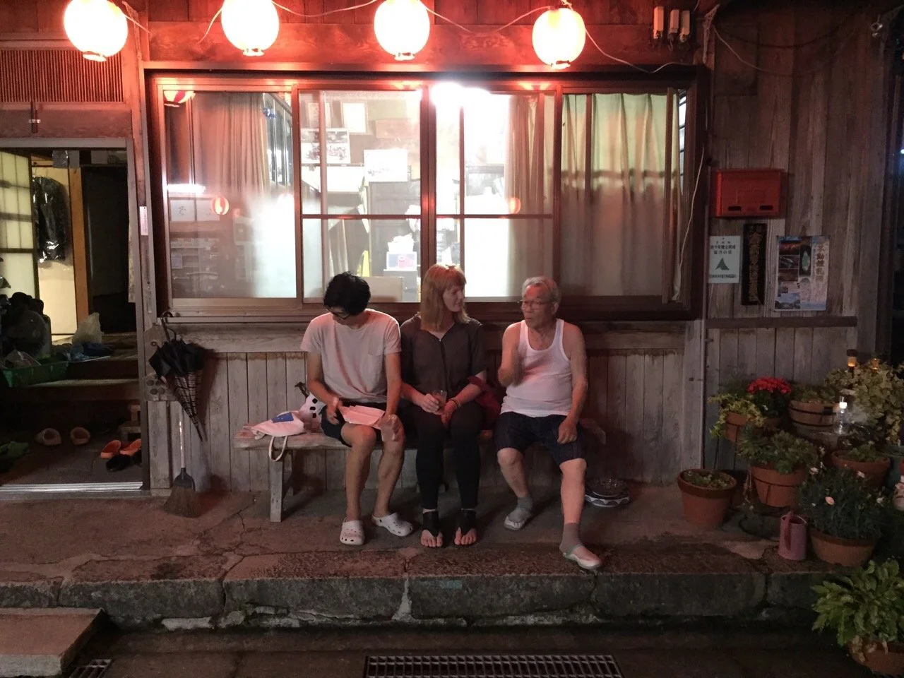 Three people sitting on a wooden bench outside a building at night, illuminated by red lanterns overhead. The person on the left is a young woman with short dark hair, wearing a white shirt and shorts. The middle person is an older woman with light h