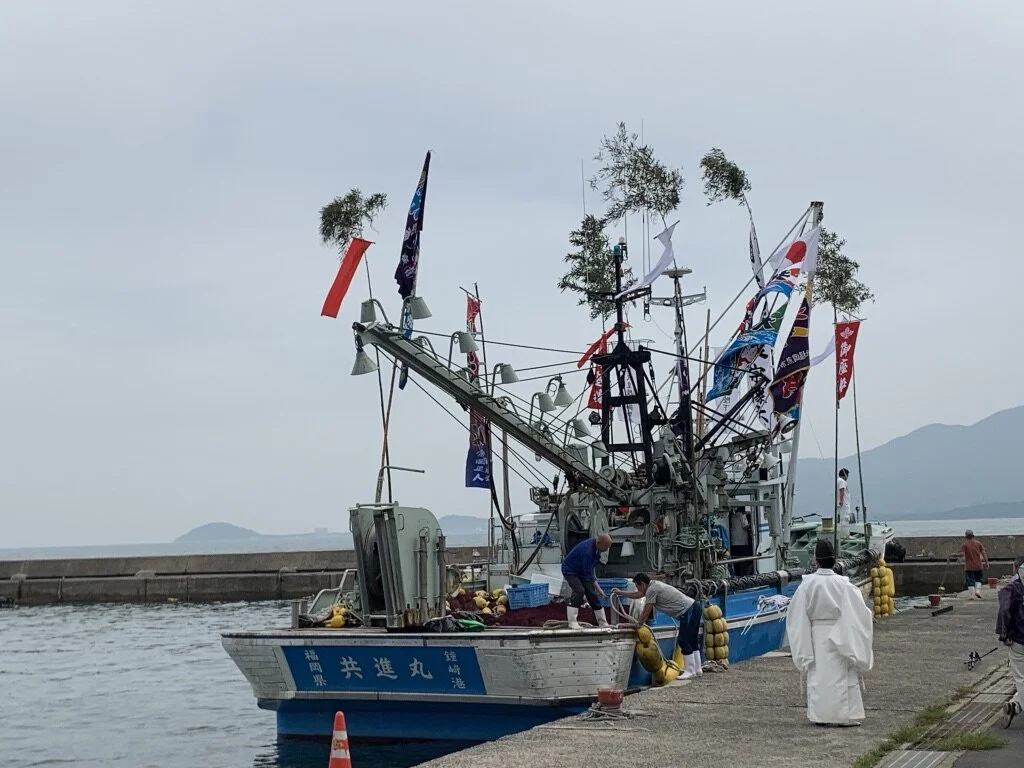 A fishing boat docked at a harbor with several people on board and on the dock, some handling fishing gear. The boat has multiple flags, including Japanese flags, and trees grow on tall poles above it. The background features a light overcast sky and