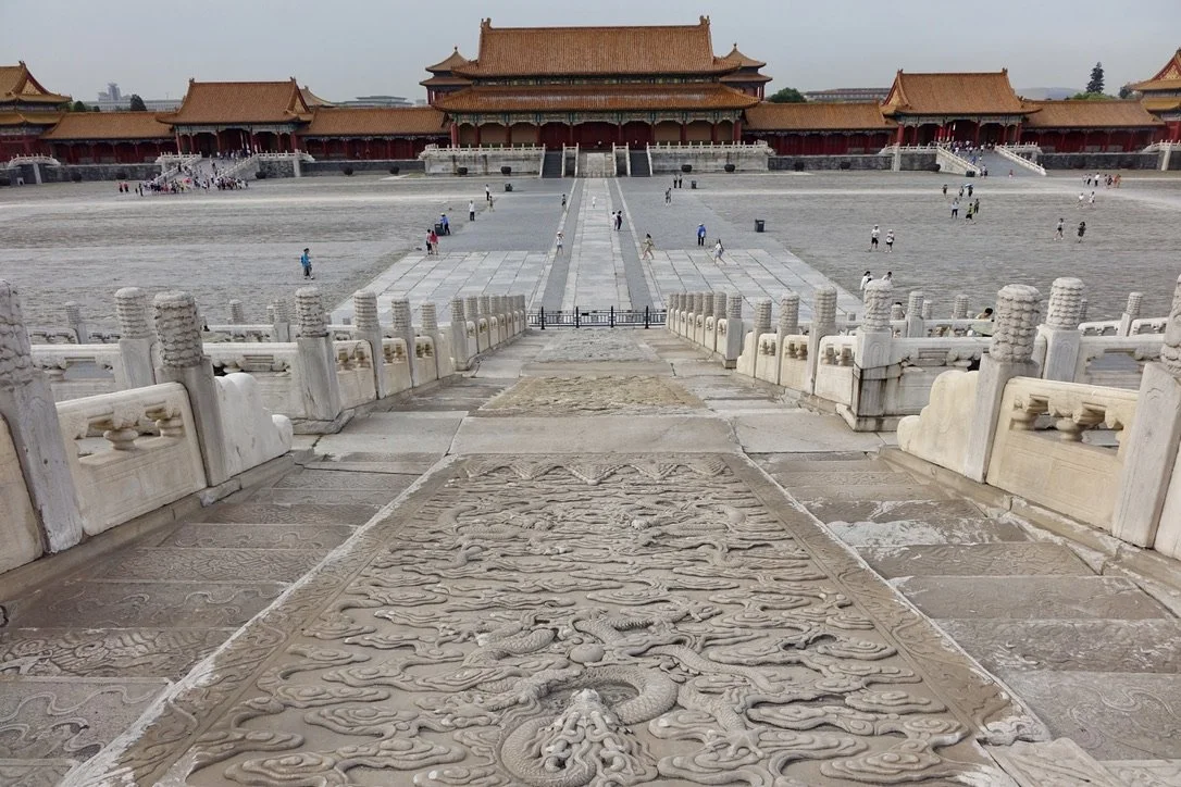 View of the Forbidden City in Beijing, China, featuring a stone dragon engraved pathway with traditional Chinese architecture and red-tiled roofs in the background, visitors walking around the courtyard.