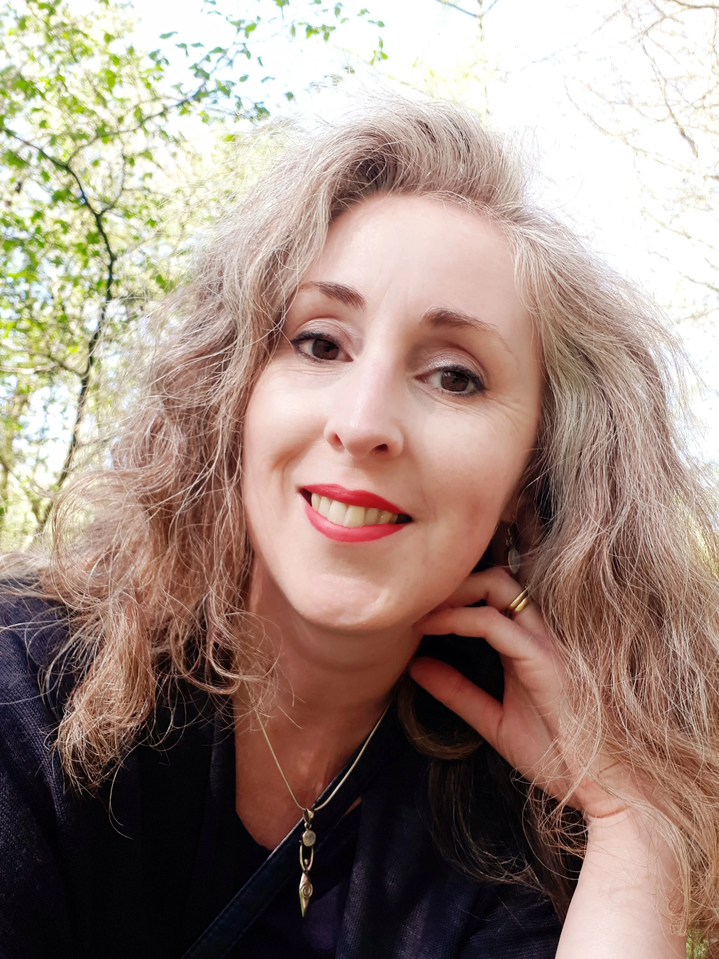 Close-up of a smiling woman with curly red-brown hair, wearing makeup, earrings, and a dark top, outdoors with green leaves and sunlight in the background.