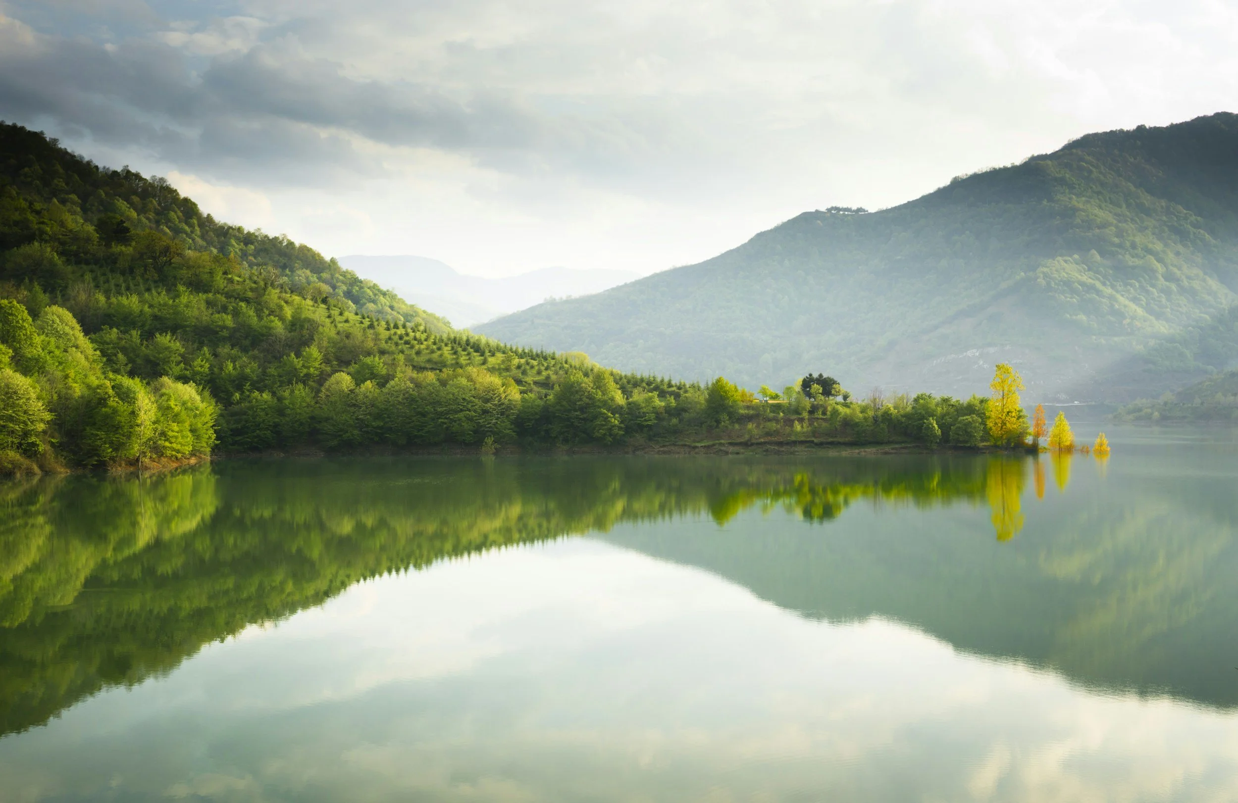 A serene landscape with green mountains reflecting in a calm body of water on a cloudy day.