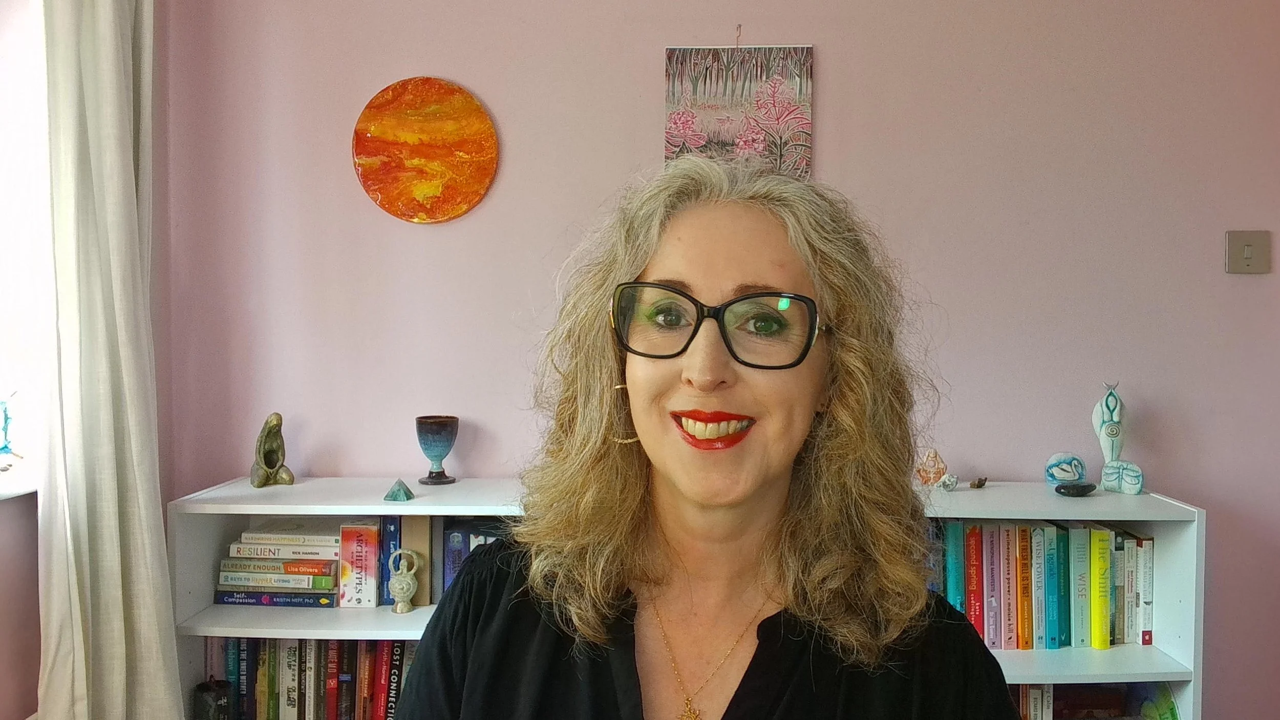 A woman with curly blonde hair, wearing black glasses and red lipstick, smiling in front of a pink wall with colorful artwork and a white bookshelf filled with books and decorative objects.