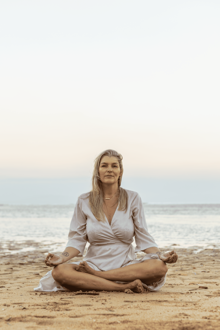 Woman sitting cross-legged on a sandy beach, practicing meditation with a calm expression, near the ocean with a clear sky.