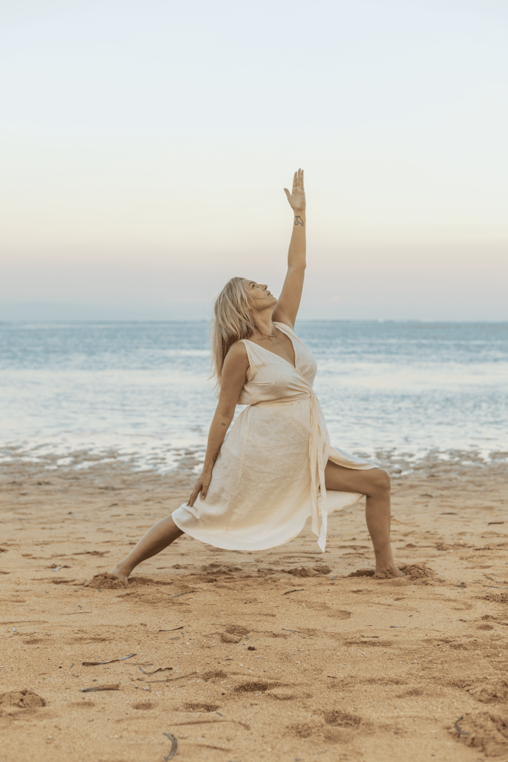 A woman practicing yoga on the beach during sunset, performing a warrior pose with her right arm extended upward and her left hand on her thigh, wearing a light-colored dress.