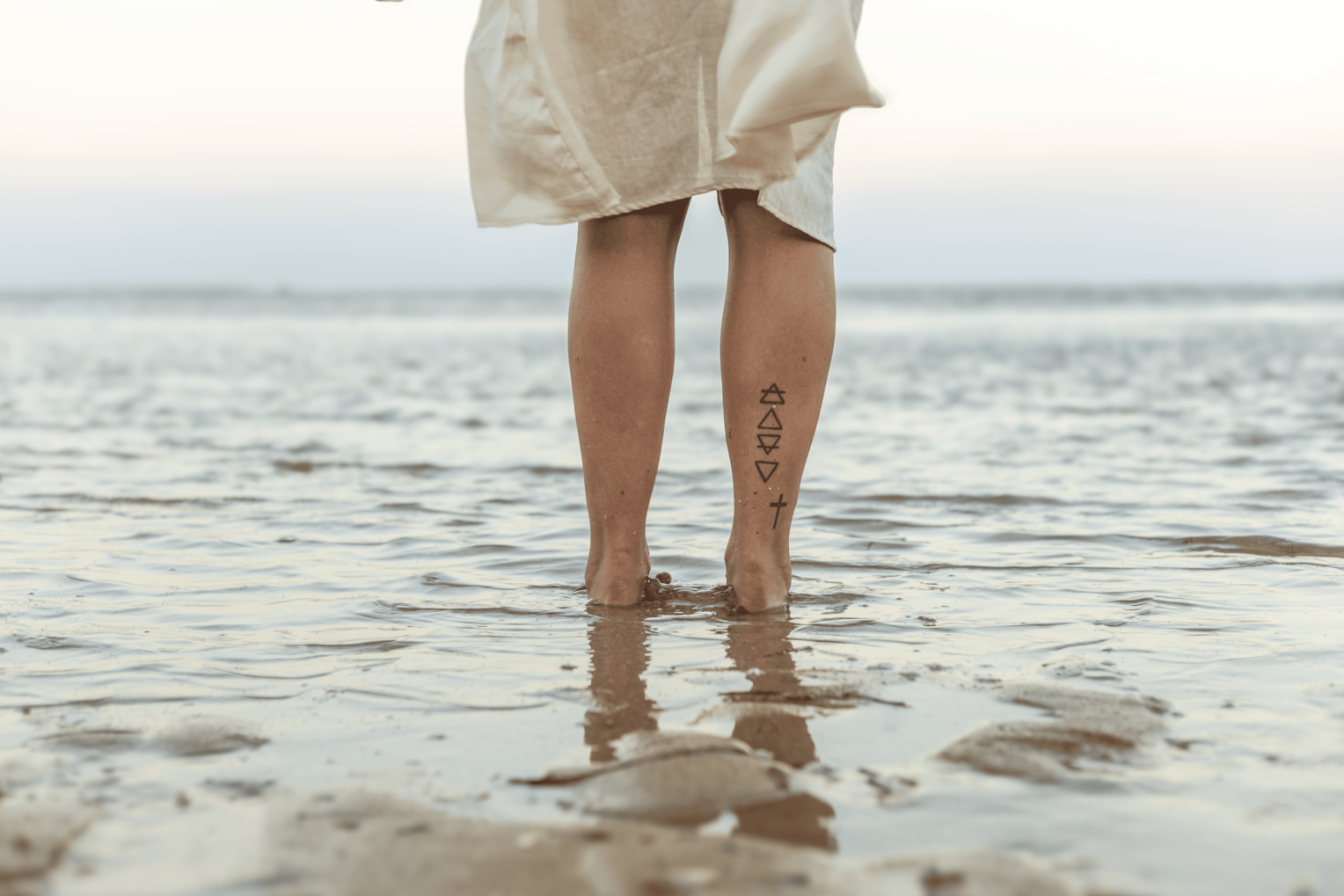 Person standing in shallow water at the beach, wearing a beige dress, showing tattoos on their calves.