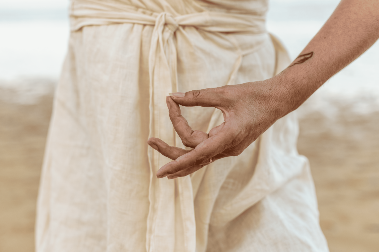 Close-up of a person's hand performing a yoga mudra gesture with the thumb and index finger touching, outdoors with a beige dress in the background.