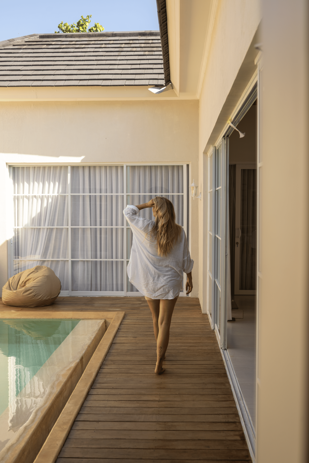 A woman with blonde hair wearing an oversized white shirt walking barefoot along a wooden poolside patio. The scene is set outside a house with sliding glass doors, a bean bag chair, and a swimming pool, with a clear blue sky overhead.