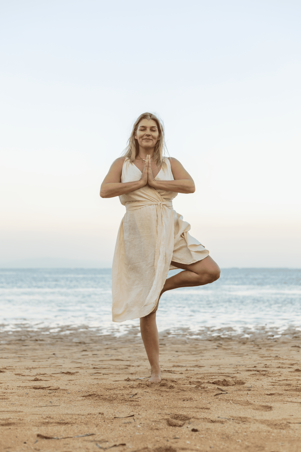 A woman practicing yoga on a beach during sunset, standing on one leg with hands in prayer position and smiling.