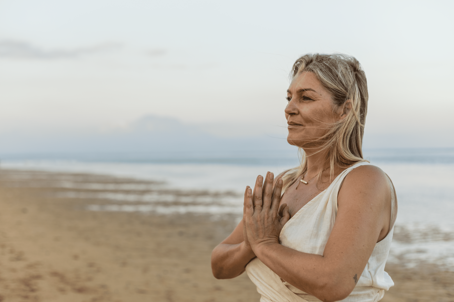 A woman practicing yoga or meditation on a beach, standing with her hands pressed together in prayer pose, peaceful expression on her face, with the ocean and cloudy sky in the background.