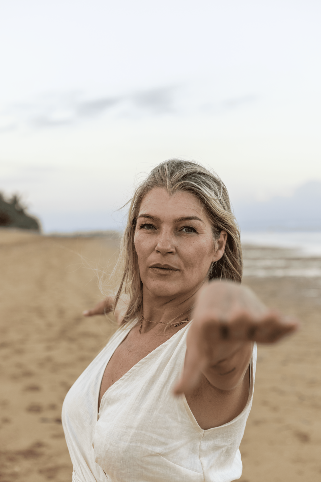 A woman on a beach reaching out towards the camera with her hand, light-colored hair, wearing a white top, ocean and cloudy sky in the background.