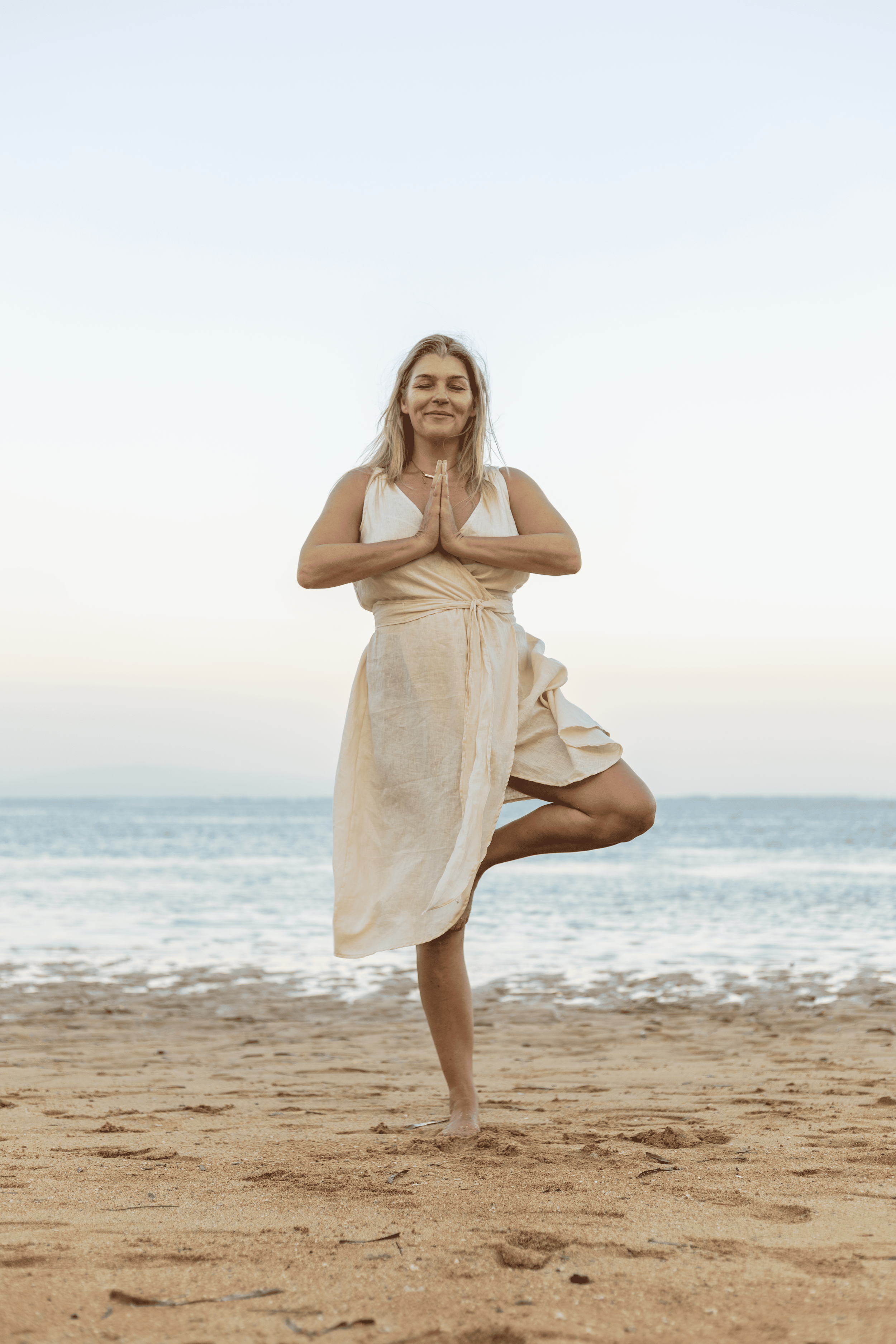 A woman practicing yoga on the beach, standing on one leg with hands together in prayer position, wearing a light-colored dress, with the ocean and sky in the background.