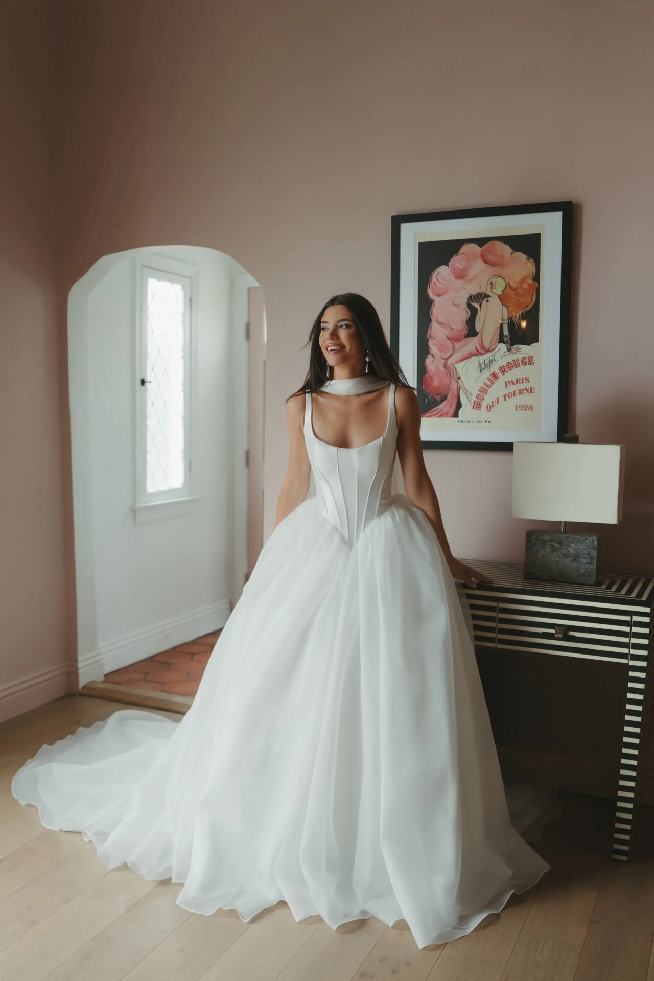 A woman in a white wedding dress standing outdoors on a brick and stone patio, with potted plants and glass-paneled doors in the background, facing away and to the left.