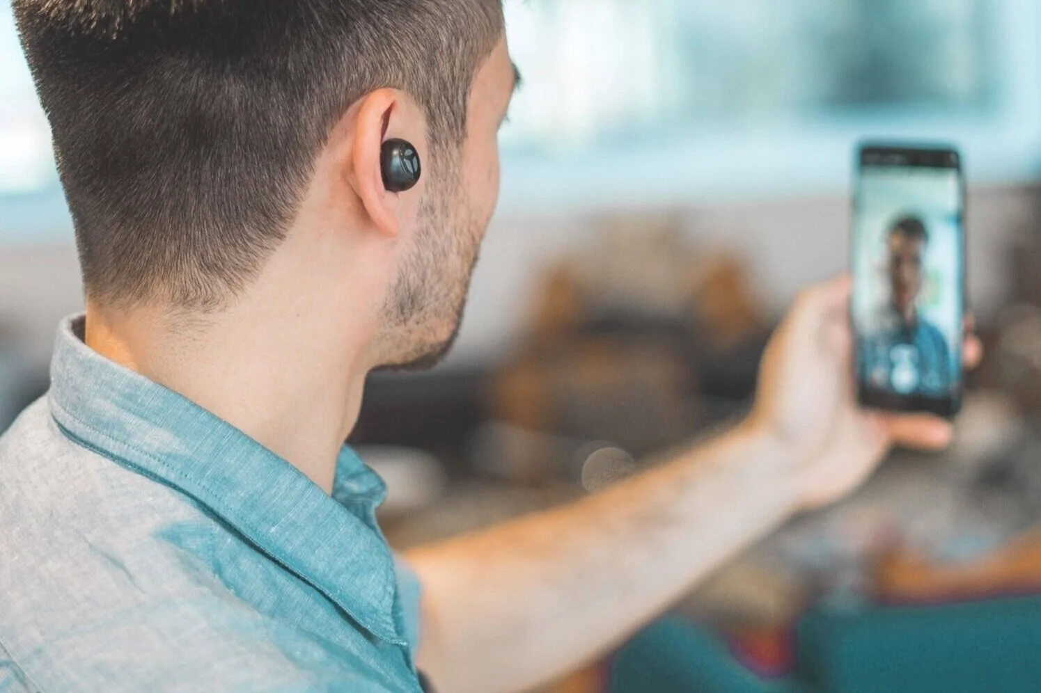 En person iført en lyseblå skjorte tager en video selfie med en smartphone. Personen har en trådløs øreprop i det ene øre.