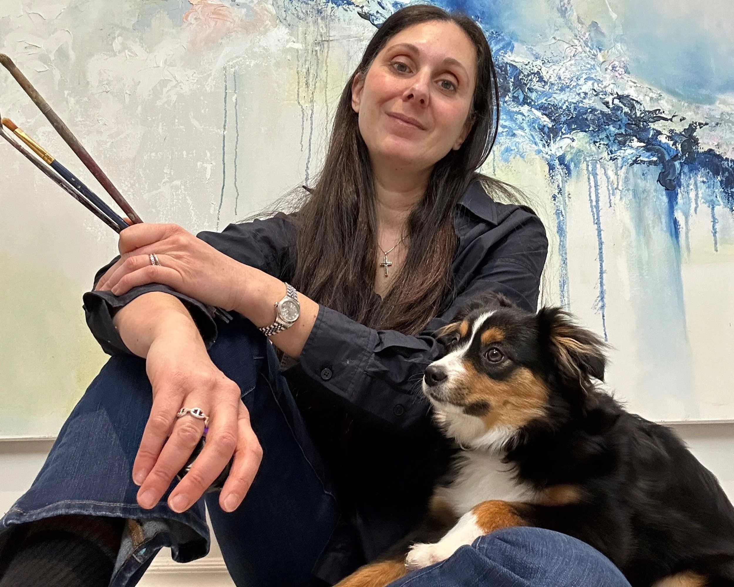 A woman with long brown hair wearing a black shirt and jewelry, sitting with a black, white, and brown puppy with floppy ears in front of a colorful abstract painting.