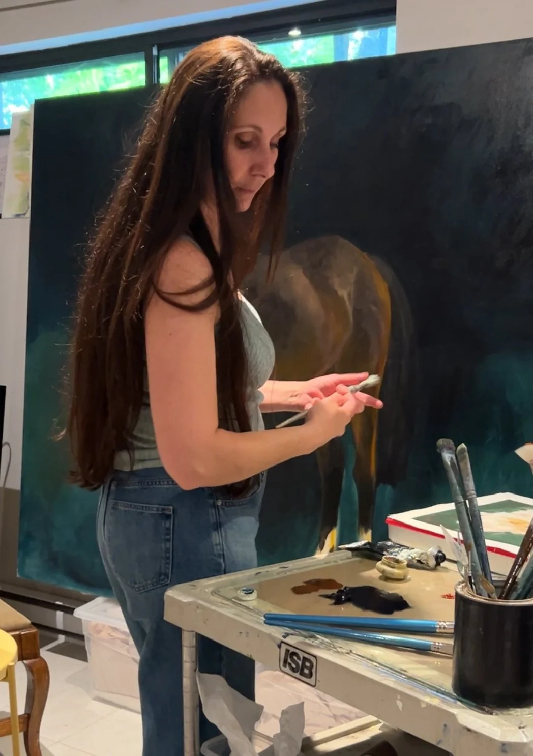 A woman with long brown hair wearing a gray tank top and jeans stands at an artist's workstation, holding a paintbrush and palette, next to a large abstract painting of an elephant.