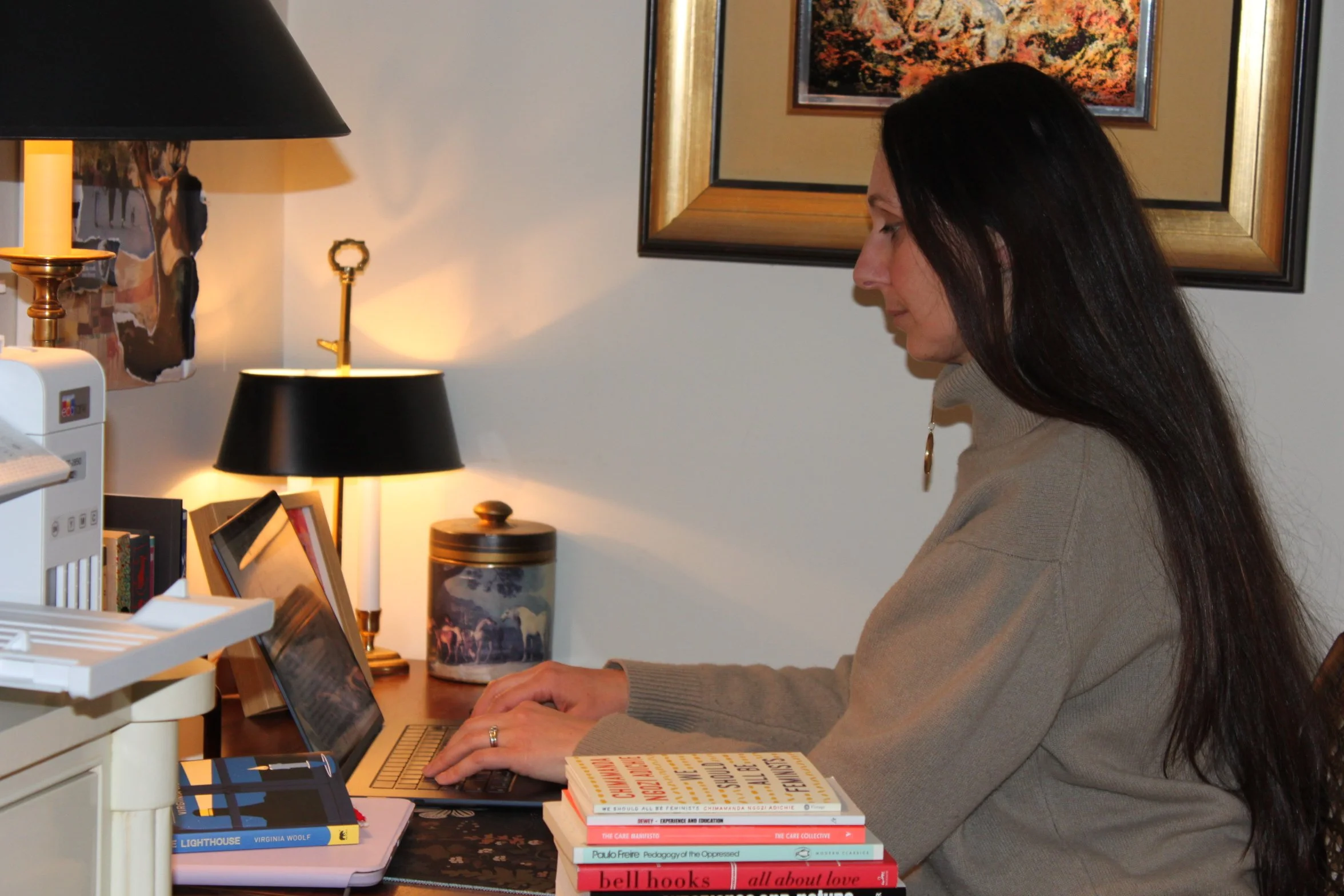A woman with long dark hair wearing a beige turtleneck sweater is sitting at a desk, using a laptop in a cozy, warmly lit room. There are books, a black-lamp, a decorative jar, and framed pictures on the wall behind her.