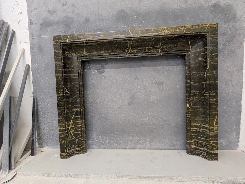 Marble fireplace mantel with a dark base color and yellowish veins, leaning against a gray textured wall.