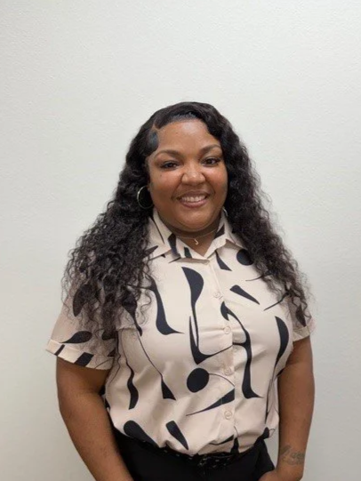 A woman with glasses and shoulder-length dark hair standing in front of a brick wall, smiling at the camera.